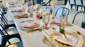 Una lunga tavola da pranzo imbandita con eleganti stoviglie e composizioni floreali, in una stanza spaziosa con soffitti alti e ampie finestre.