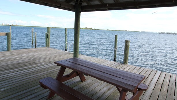 Un muelle de madera con una mesa de picnic, que se extiende sobre aguas tranquilas bajo un cielo despejado.