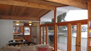L’intérieur chaleureux d’un chalet avec un plafond en bois, un coin repas comprenant une table et des chaises, et une grande porte-fenêtre coulissante donnant sur un balcon avec vue sur un lac et des bâtiments.