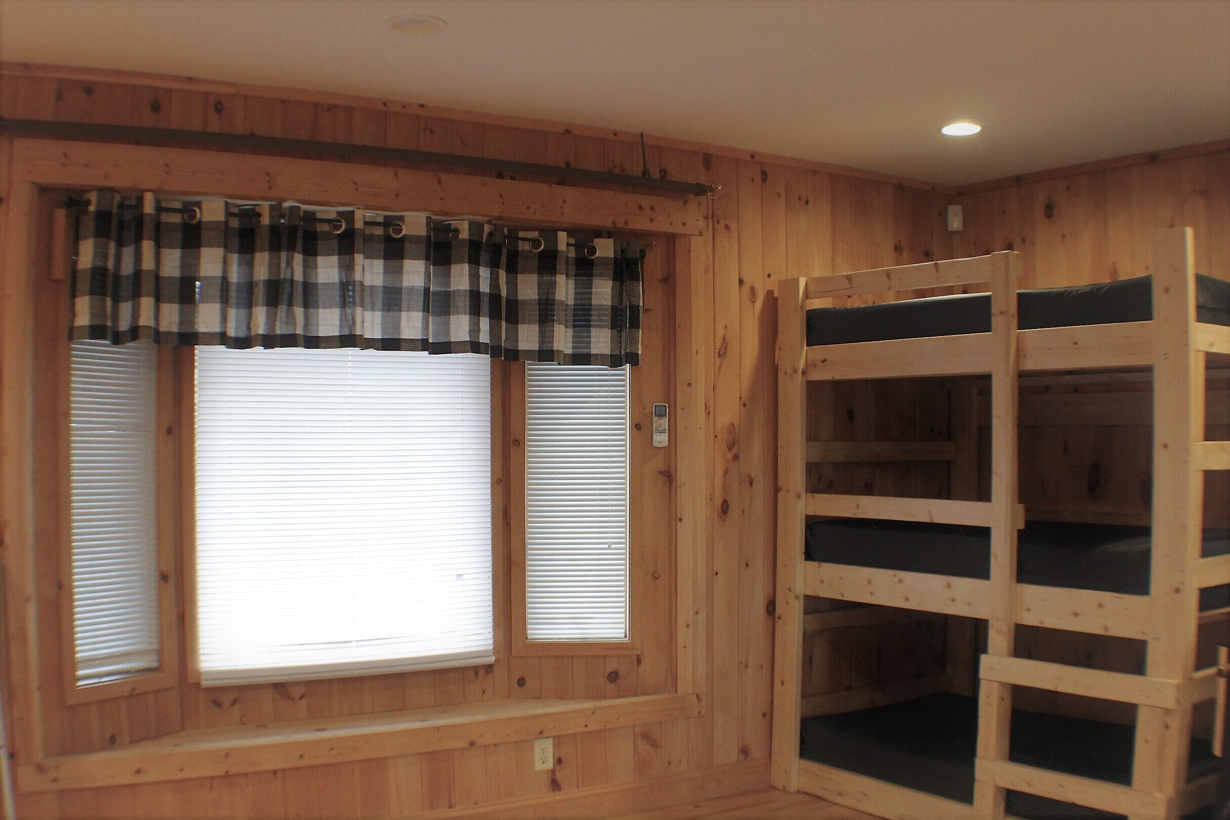 A room with wooden walls, a bunk bed, and two windows with blinds.