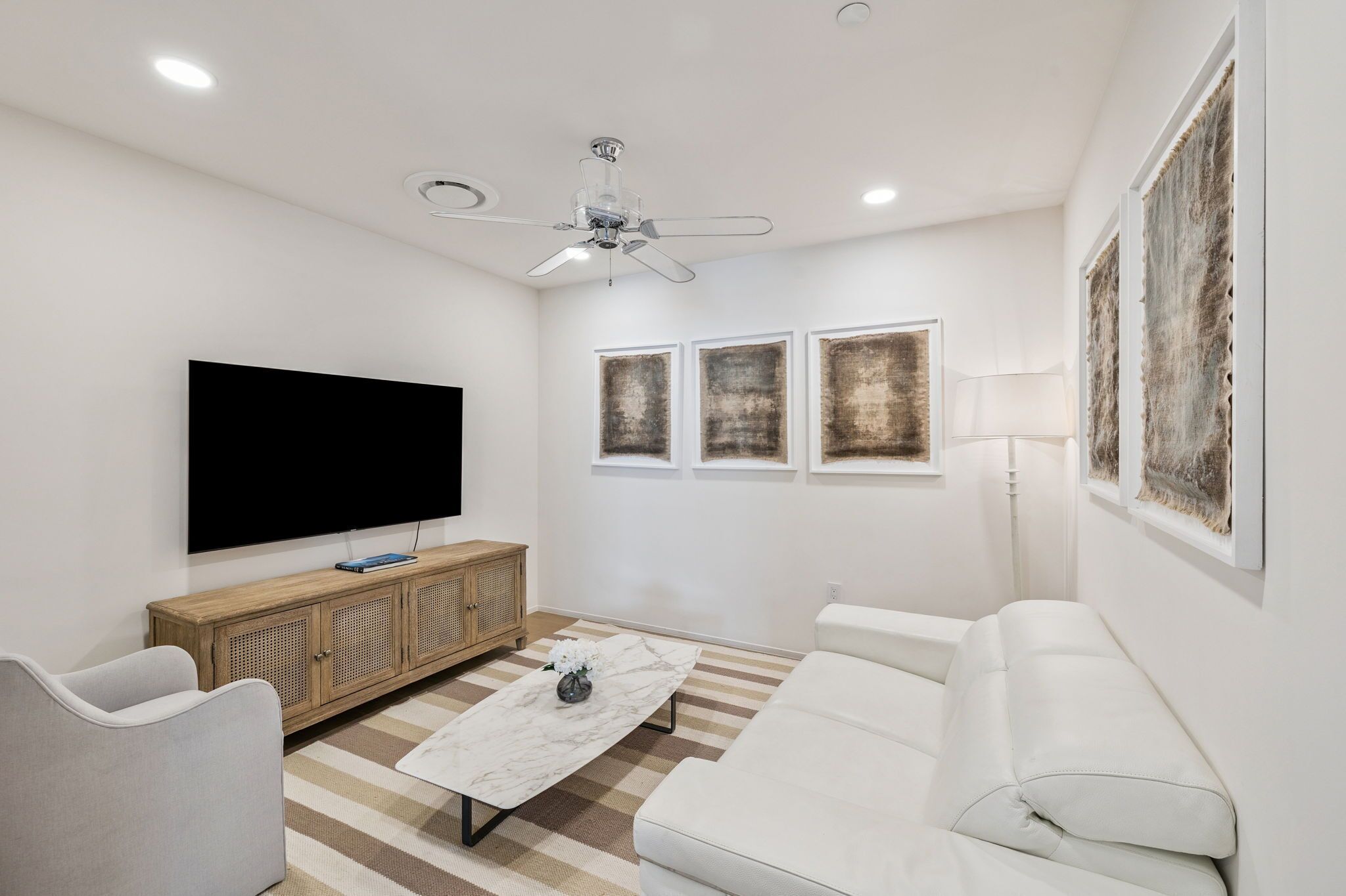 Un soggiorno moderno con una TV a schermo piatto, un mobiletto in legno, un divano bianco, un tavolino da caffè in marmo e una ventola a soffitto.