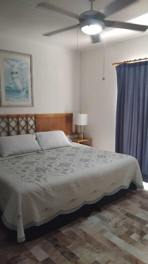 A bedroom with a bed, a nightstand, a ceiling fan, a lamp, a painting, and a window with curtains. - Sleep by the Blue Paradise (Cancun)
