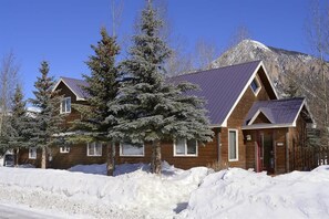 A wooden cabin with a sloped roof, surrounded by snow-covered ground and trees, with a mountain in the background.