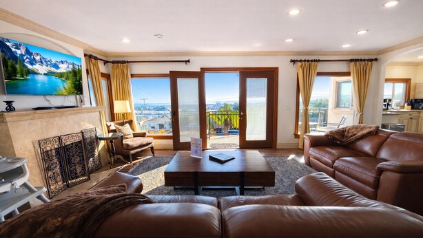 Ein geräumiges Wohnzimmer mit einem großen Sofa, einem Couchtisch und einem Kamin. Große Fenster mit Vorhängen, ein Balkon und ein sichtbarer Küchenbereich sind ebenfalls vorhanden.