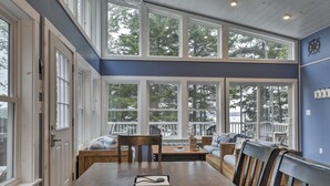 A dining area with a wooden table, chairs, and a view of the outdoors through large windows. - Enjoy the quiet, the calm, the sunset and the deck! (Cushing)