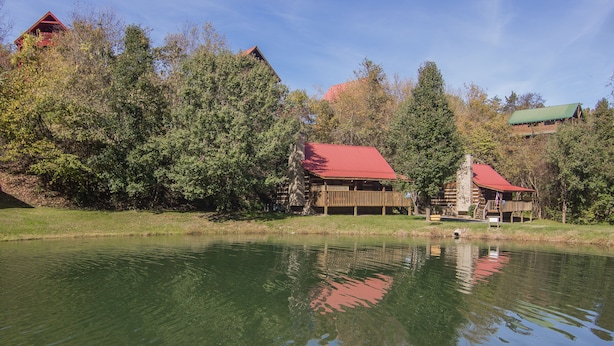 A rustic cabin with a red roof by a calm lake, surrounded by dense trees and a clear sky.