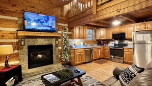 A cozy cabin interior with a stone fireplace, wooden ceiling, a television mounted above it, a Christmas tree, a sofa, and a kitchen area with wooden cabinets and appliances.