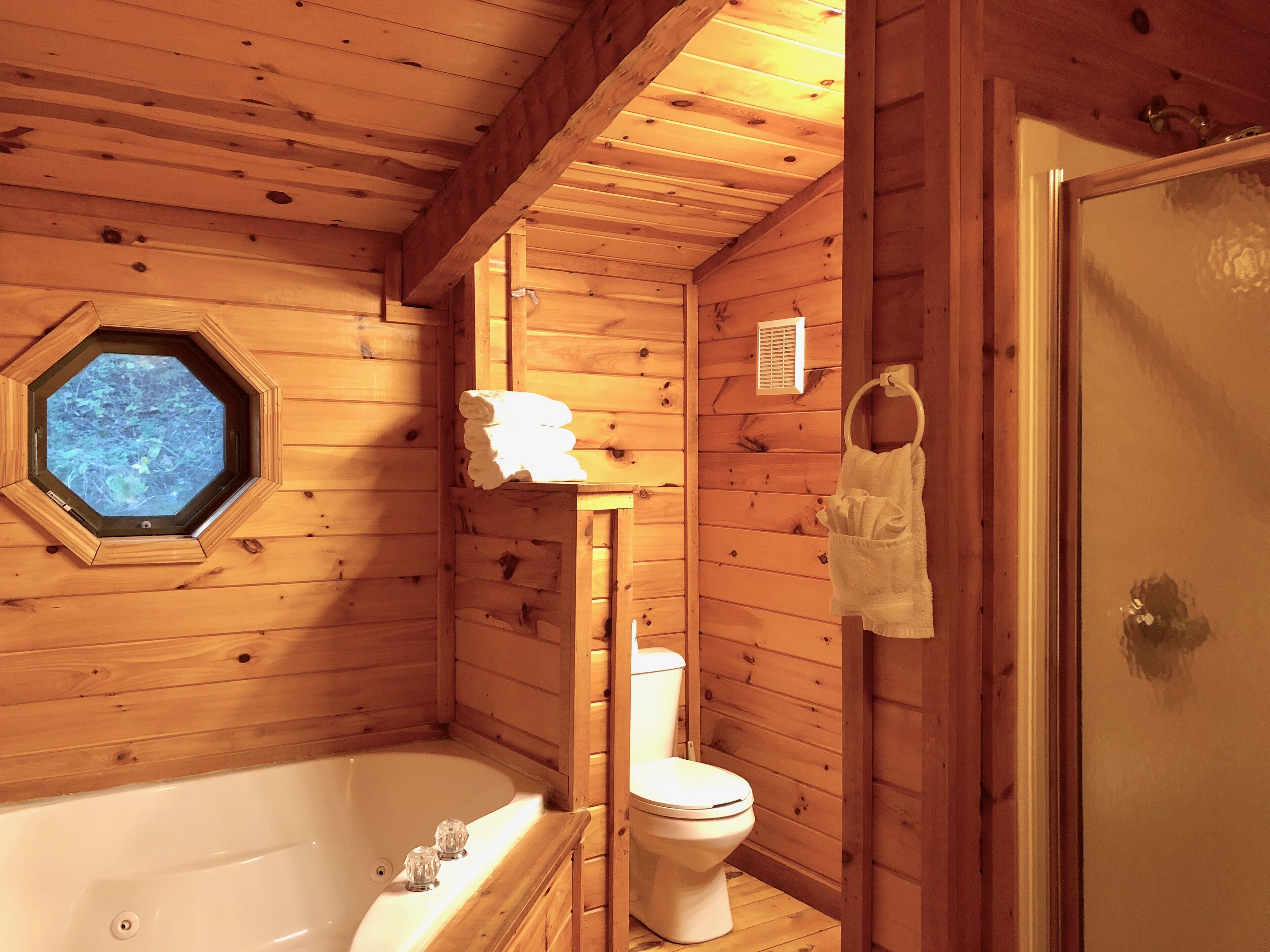 A wooden cabin bathroom with a bathtub, toilet, and a window with a view of trees.