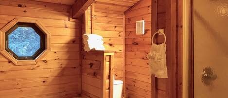 A wooden cabin bathroom with a bathtub, toilet, and a window with a view of trees.