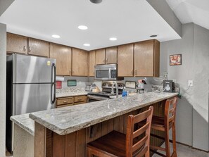 A kitchen with wooden cabinets, stainless steel appliances, a marble countertop, and a built-in island with bar stools. - Ski-In/Out Giant Steps Condo | Sleeps 12 (Brian Head)