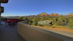 A balcony with red chairs and a table, overlooking a scenic view of red rock formations and a town below.