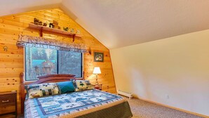Un dormitorio acogedor con techo inclinado de madera, un ventanal con vistas a una cabaña, una cama con colcha, una mesita de noche y un aparador.