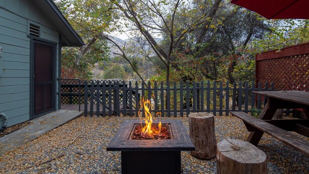A square fire pit with flames, surrounded by gravel and wooden stumps, in a backyard with a wooden fence and a picnic table. - 1 Min from Sequoia National Park Cabin for 2 with additional sleeper sofa (Three Rivers)