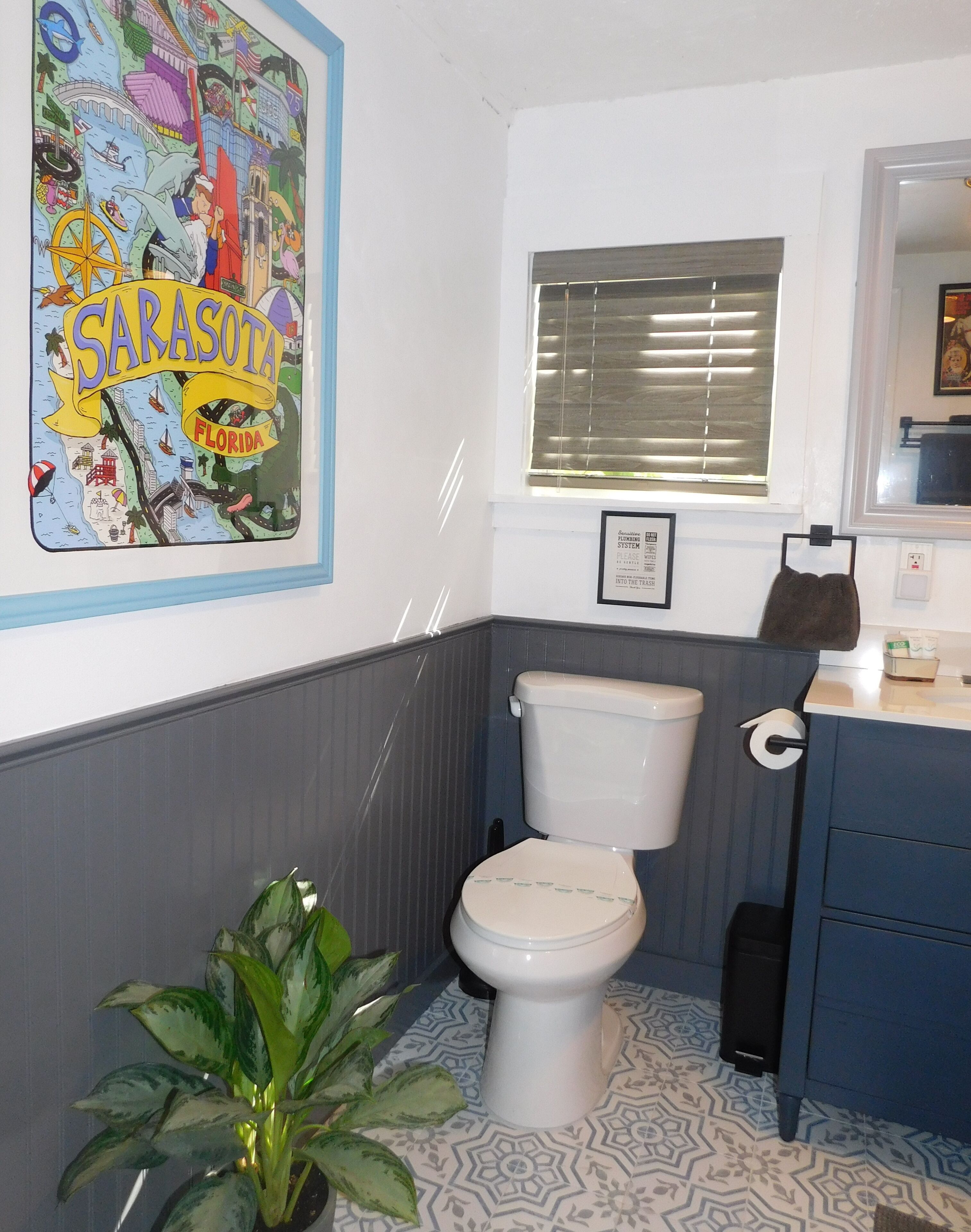 A bathroom with a toilet, a sink, a mirror, a window with blinds, a framed certificate, and a potted plant.