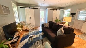 A living room with a sofa, coffee table, and a TV. There are two doors with curtains, a window, and a lamp.