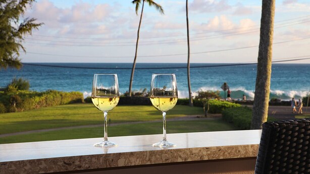 Dos copas de vino blanco en un balcón con vista a una playa, palmeras y el océano.