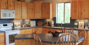 A kitchen with wooden cabinets, a microwave, a stove, a dining table with chairs, and a fruit basket.