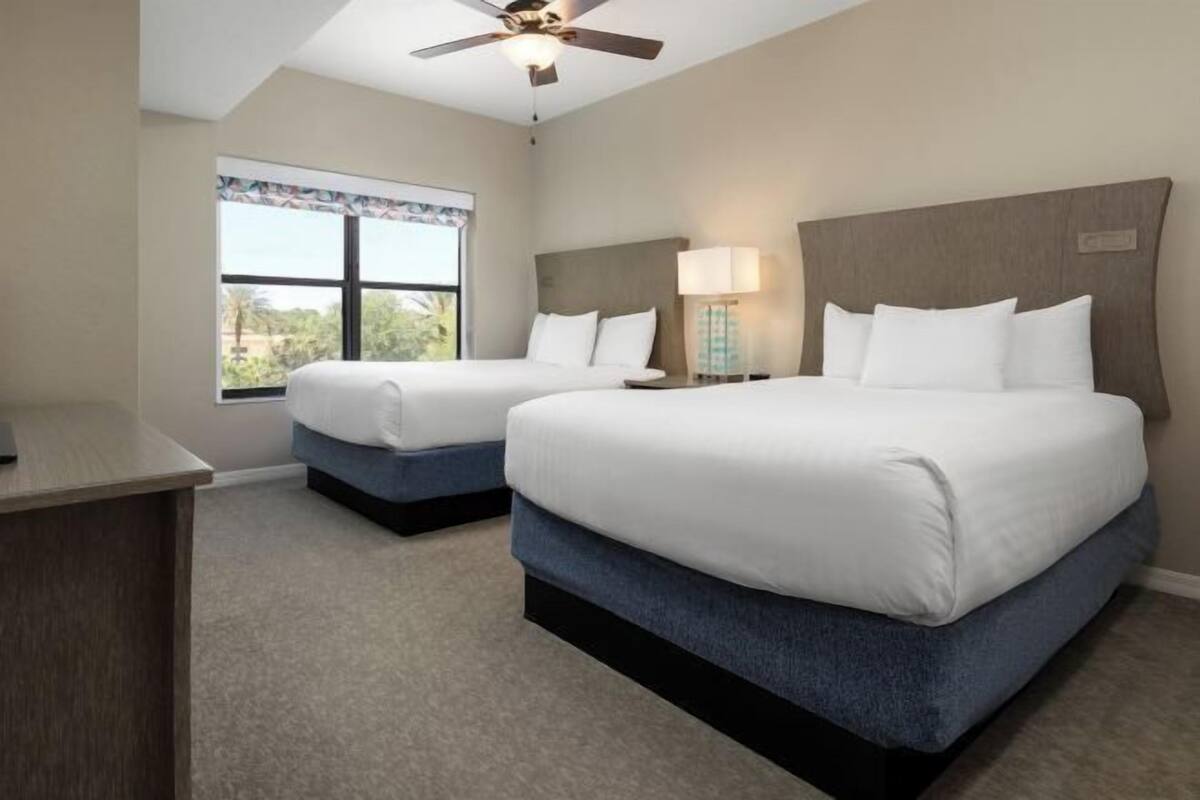 A hotel room with two beds, a desk, a ceiling fan, a window with a view, and a lamp.