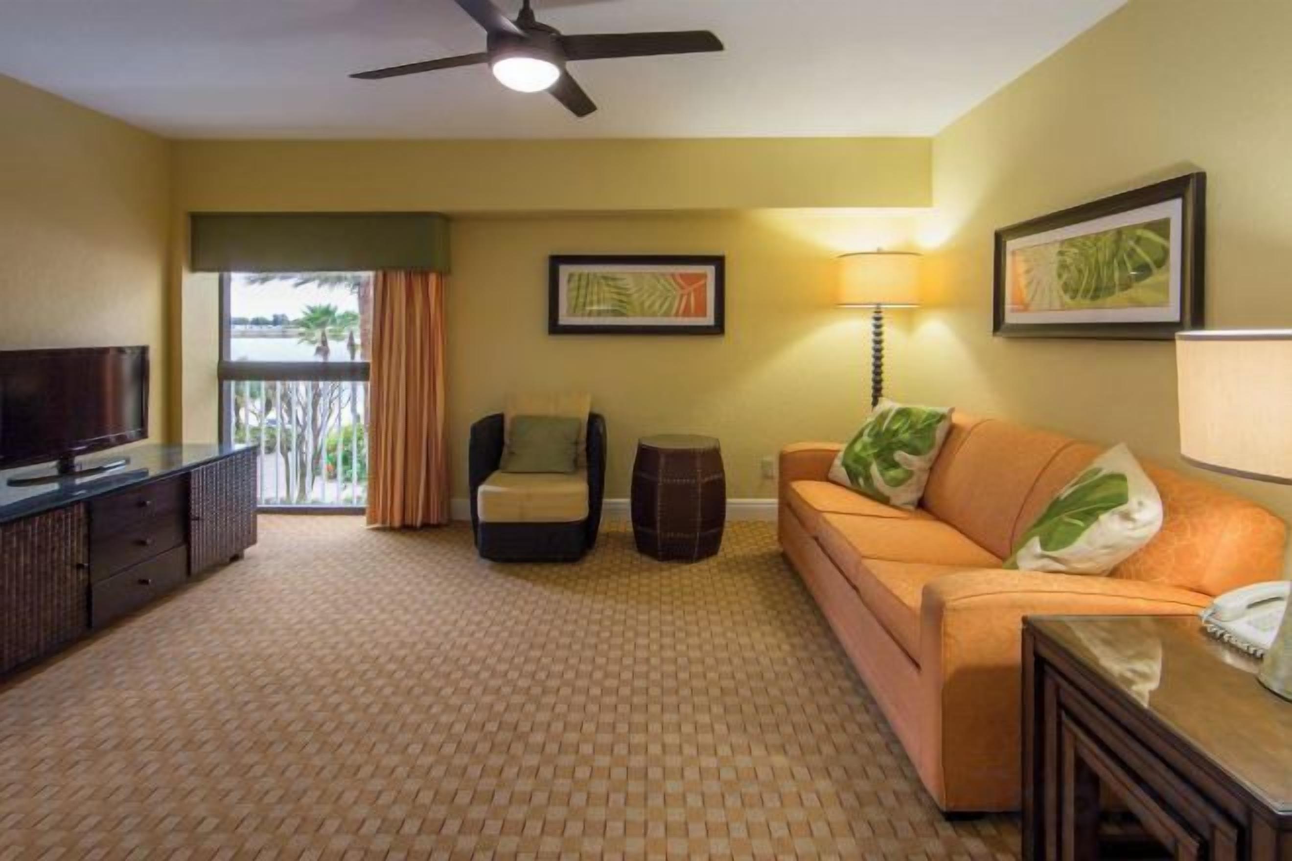 A hotel room with a television, a sofa, a chair, a lamp, and a ceiling fan.