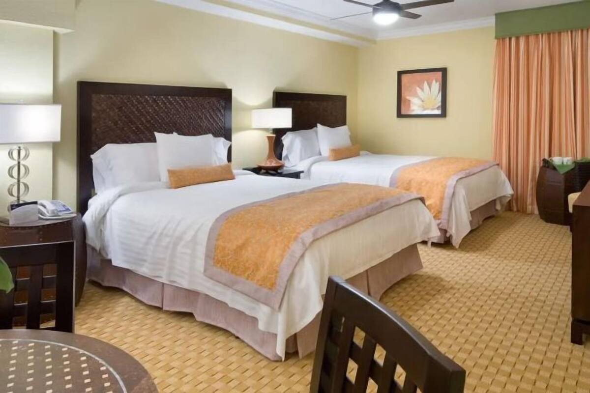 A hotel room with two beds, a desk, a chair, a lamp, and a ceiling fan.