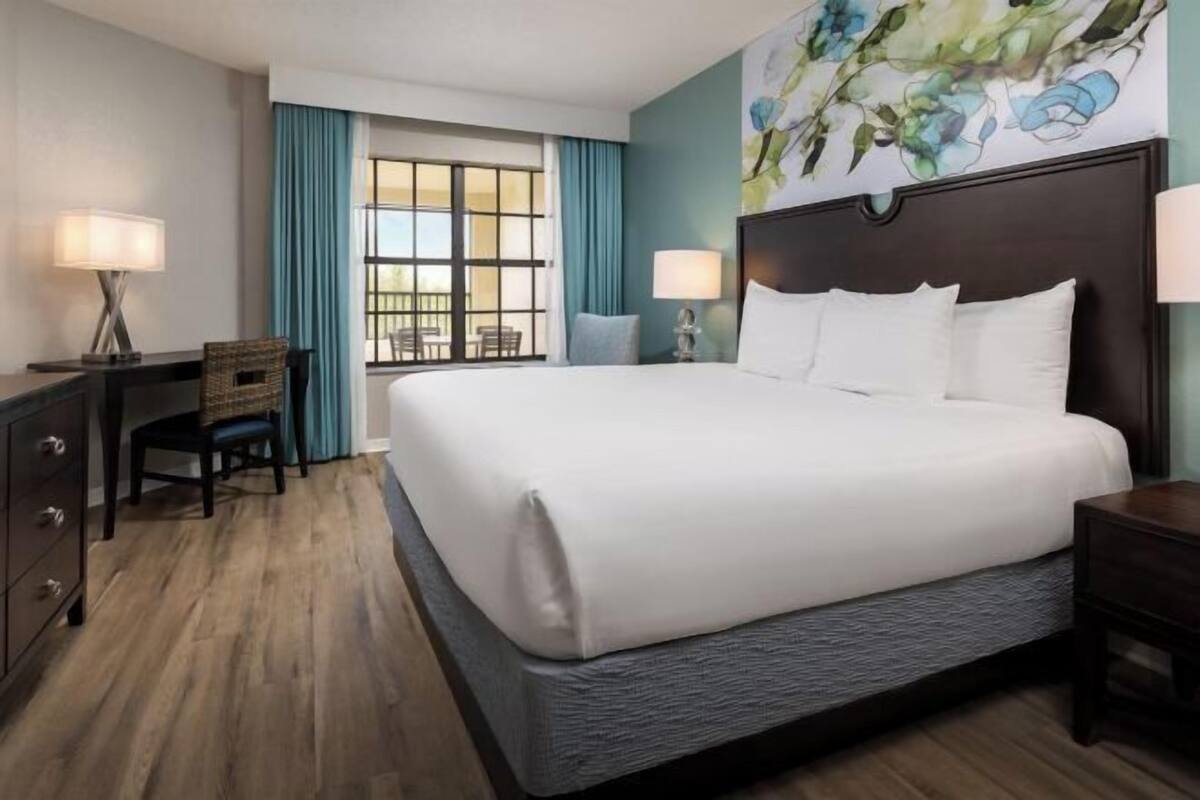 A hotel room with a large bed, a desk, a chair, a window with curtains, and a painting on the wall.