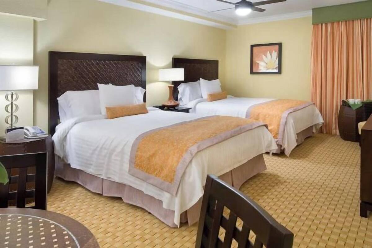 A hotel room with two beds, a desk with a lamp, a chair, a ceiling fan, and a framed picture on the wall.