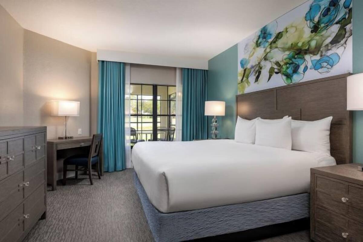 A hotel room with a bed, desk, chair, dresser, and a large floral artwork on the wall.