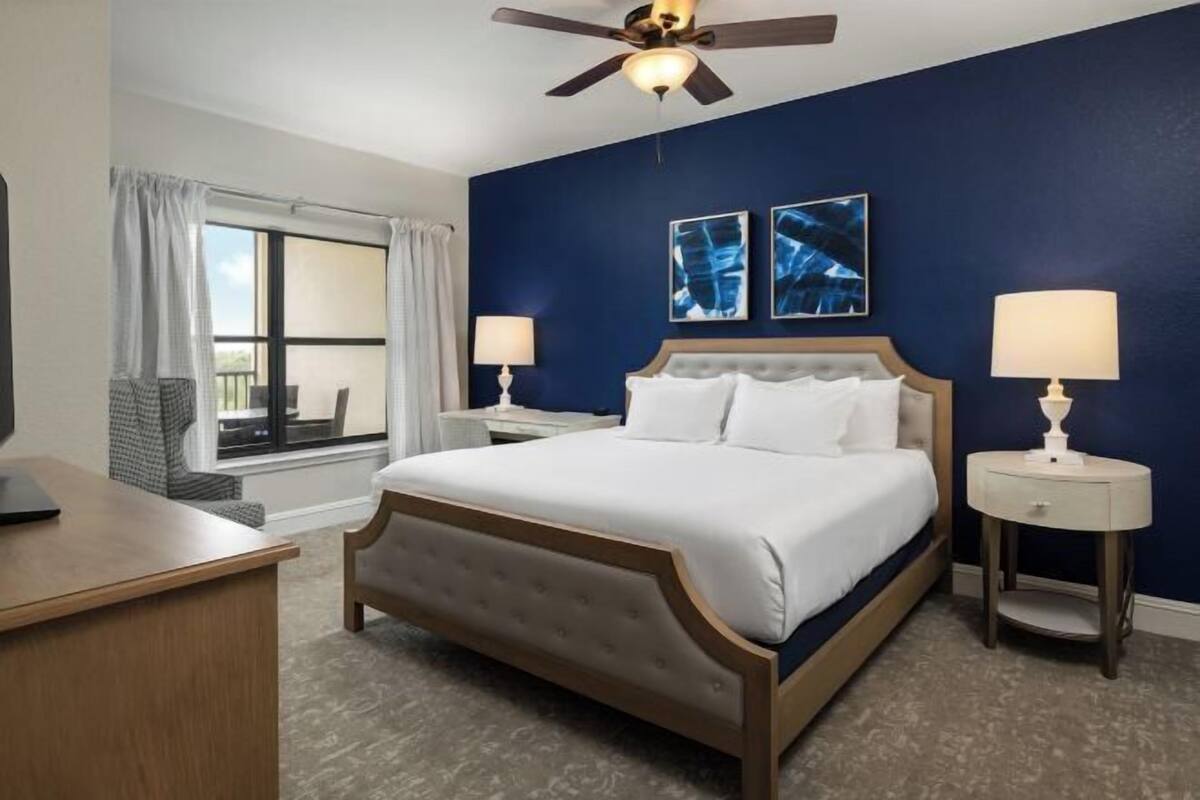 A hotel room with a bed, two bedside tables, a desk, a ceiling fan, and artwork on the wall.
