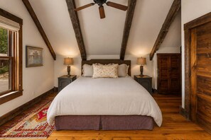 A bedroom with a large bed, wooden beams, two bedside tables with lamps, a window, and a door. - The Restoration (Crested Butte)