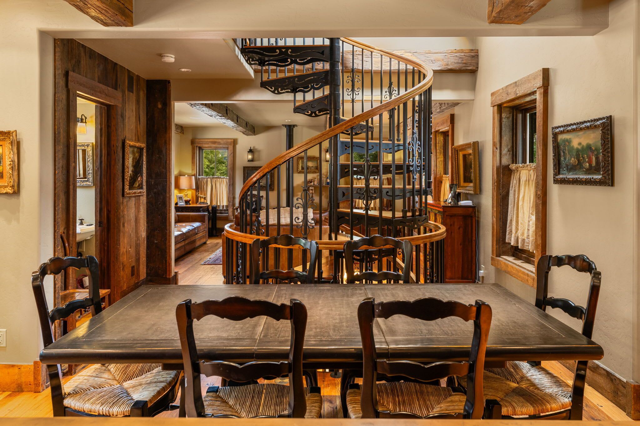 A dining area with a large wooden table and chairs, a spiral staircase, and a view into another room with a window.