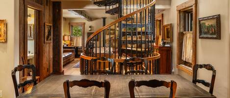 A dining area with a large wooden table and chairs, a spiral staircase, and a view into another room with a window.