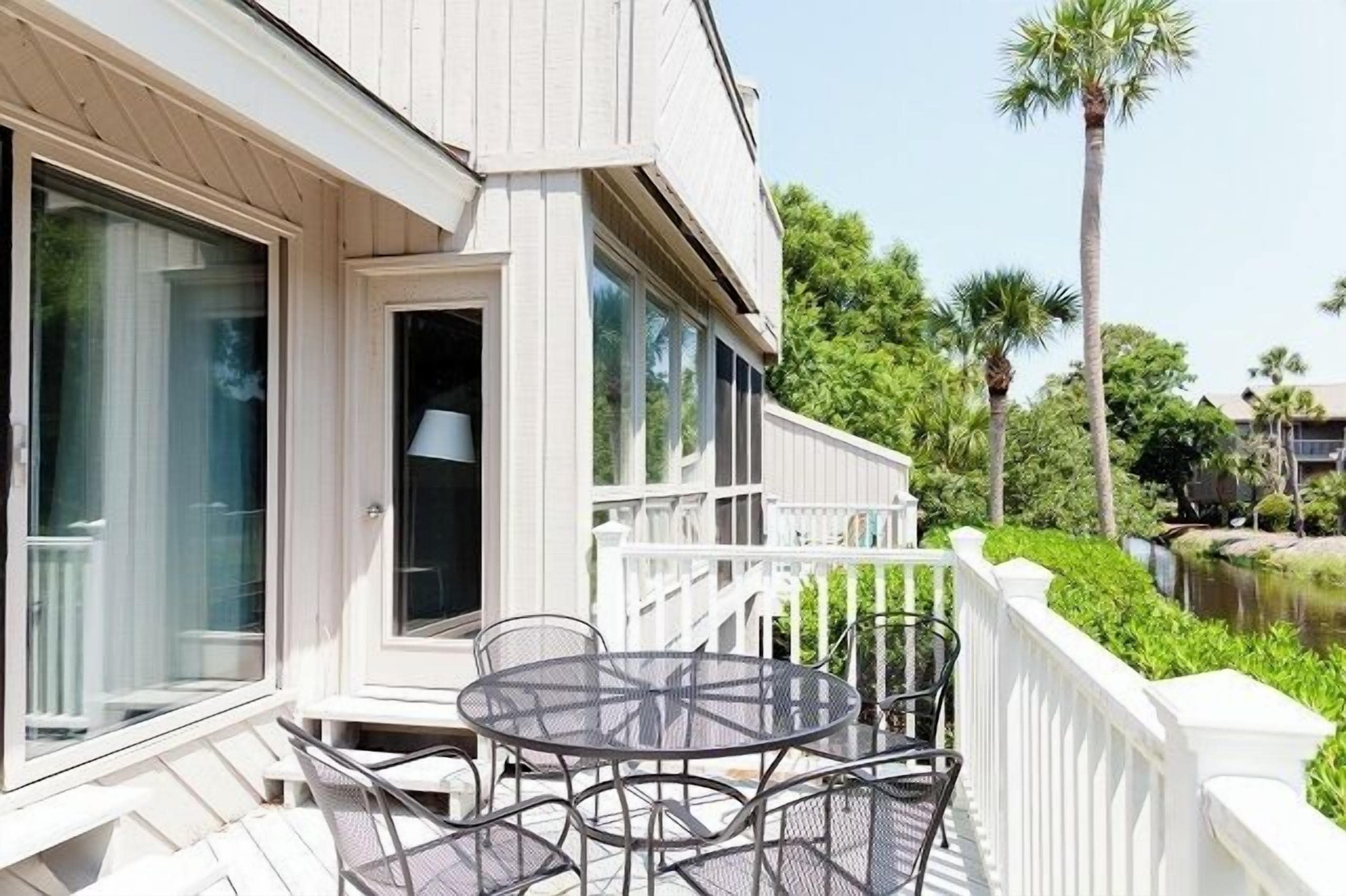 A covered outdoor area with a round table and chairs, a white railing, and a view of a water body and palm trees.