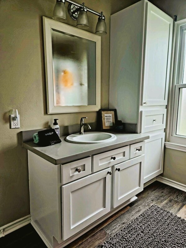 A bathroom with a white vanity, a large mirror, and a sink. There are three lights above the mirror, and a small framed picture hangs on the wall.