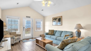 A spacious living room with a high ceiling, a ceiling fan, a large window, a sofa, a coffee table, and a lamp.