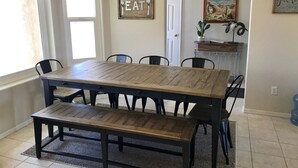 Uma sala de jantar com mesa e bancos de madeira, um candelabro e obras de arte nas paredes, incluindo uma placa com a palavra 'EAT' e uma pintura de uma paisagem desértica.