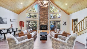 A spacious living room with a high ceiling, a stone fireplace, and large windows offering views of trees. - Empty Saddle - 6 bedrooms / 3.5 baths (Duck Creek Village)