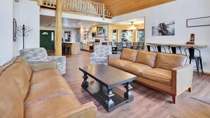 A spacious living area with a high ceiling, wooden beams, a chandelier, leather sofas, a dining table, and a kitchen in the background. - Empty Saddle - 6 bedrooms / 3.5 baths (Duck Creek Village)
