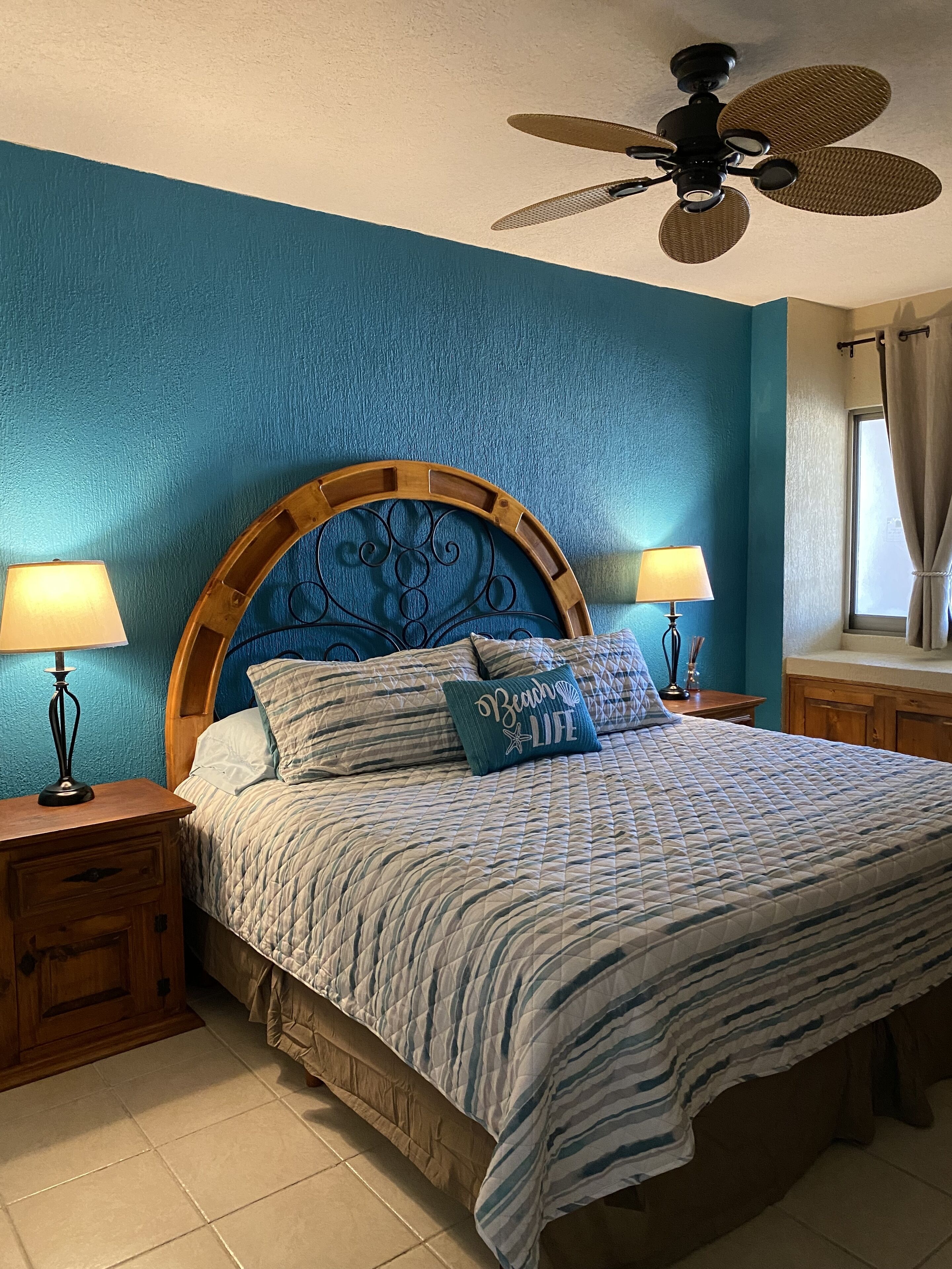 A bedroom with a large bed, two bedside tables with lamps, a ceiling fan, and a window with curtains.