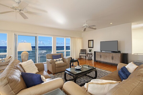 A spacious living room with a large window overlooking the ocean, a ceiling fan, a television, and a seating area with a coffee table.