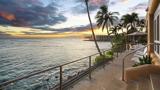 A balcony with a view of the ocean at sunset, palm trees, and a lounge area with chairs.