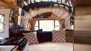 Ein kompaktes Wohnmobil-Interieur mit einer Küchenzeile an einer Seite, einem Bett mit gemusterten Bettwäsche und einer gewölbten Decke.