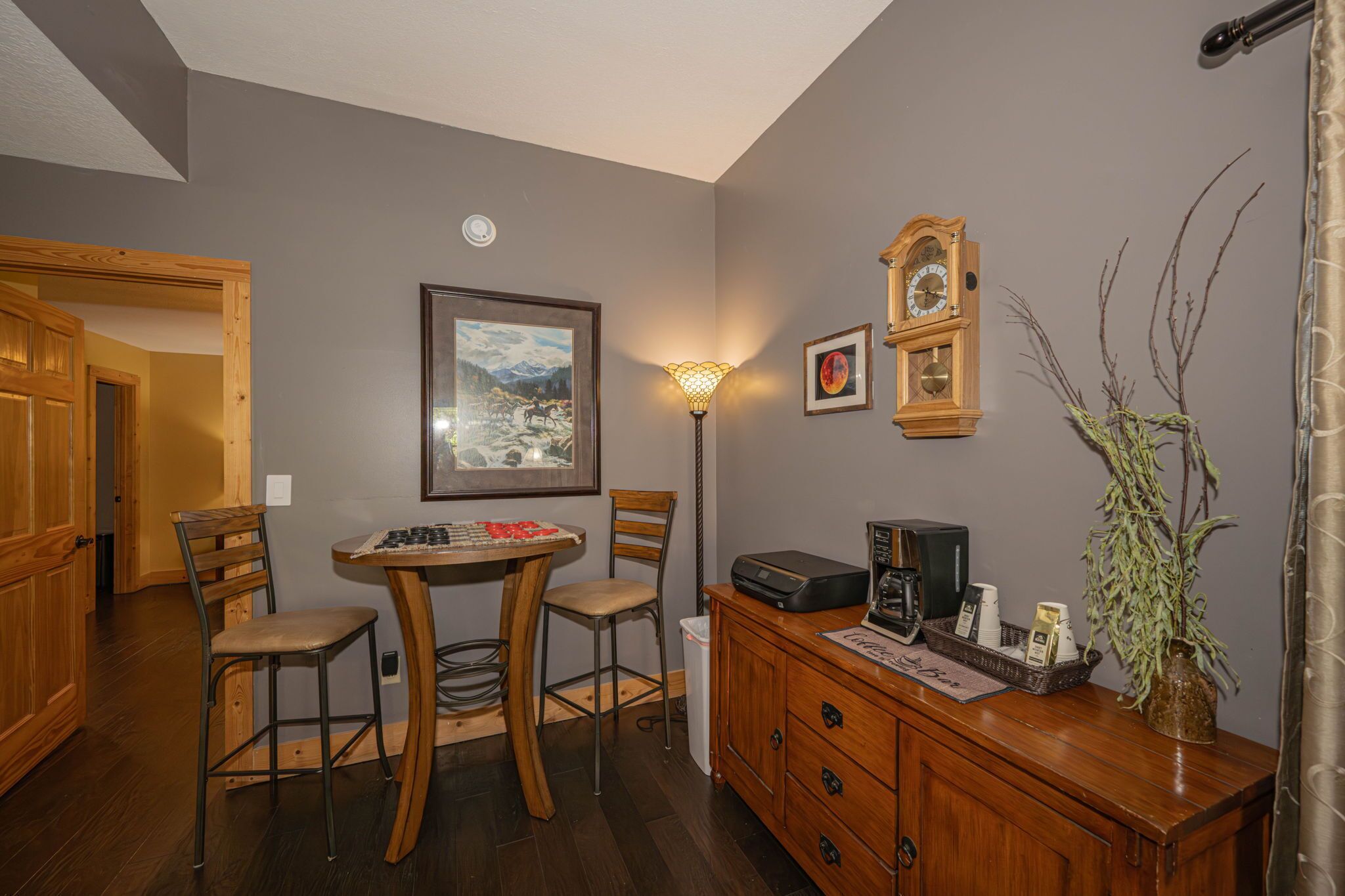 A dining area with a table, chairs, a lamp, a clock, a framed picture, and a cabinet with a vase.