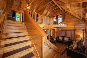 Interior de una cabaña de madera con escalera, una sala de estar con sofá y un balcón.