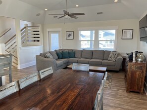 A spacious living room with a large dark wood dining table, a grey sectional sofa, and a ceiling fan.