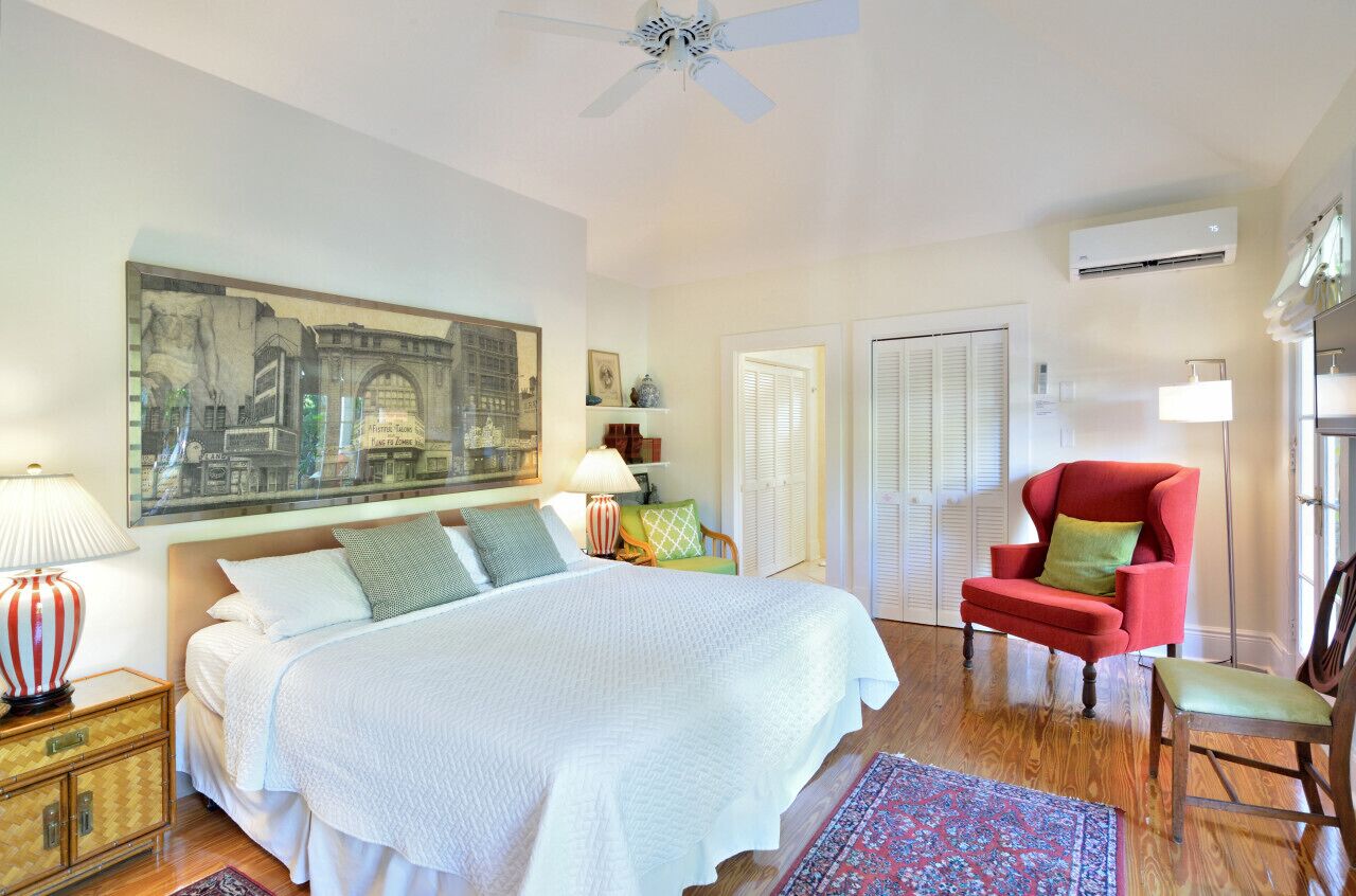 A bedroom with a large bed, a red armchair, a wooden nightstand, a floor lamp, and a ceiling fan.