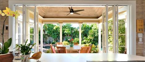 A modern kitchen with a central island, a ceiling fan, and a view of a lush garden through open doors.