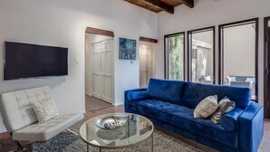 Een woonkamer met een blauwe bank, een witte fauteuil, een glazen salontafel en een flatscreen televisie aan de muur.