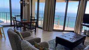 A spacious living room with a large window offering a view of the ocean, a dining area with chairs and a table, and a black leather ottoman. - DISCOUNTED RATES!   BREATHTAKING VIEWS FROM BEAUTIFUL CORNER UNIT!  3 BR/2 BA. (Orange Beach)