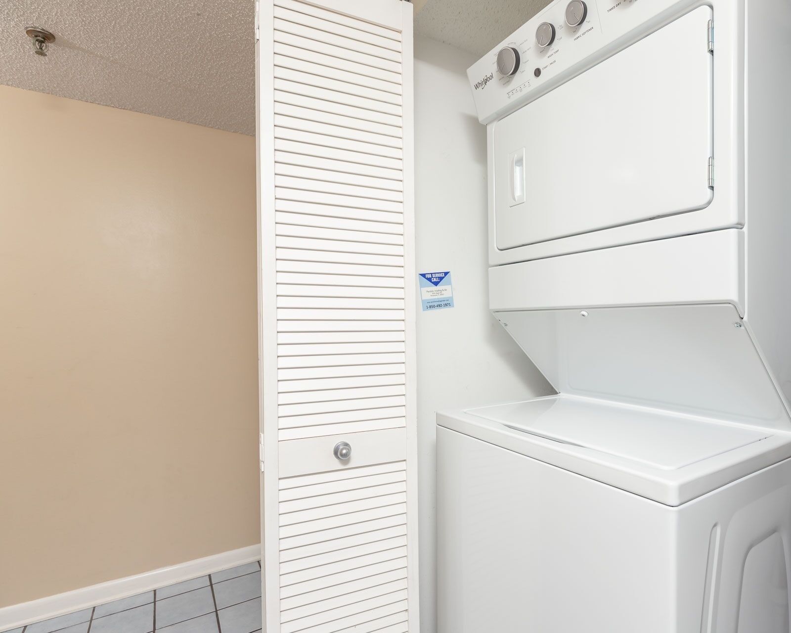 A laundry room with a front-loading washer and dryer, a closed closet door, and a service call sign on the wall.