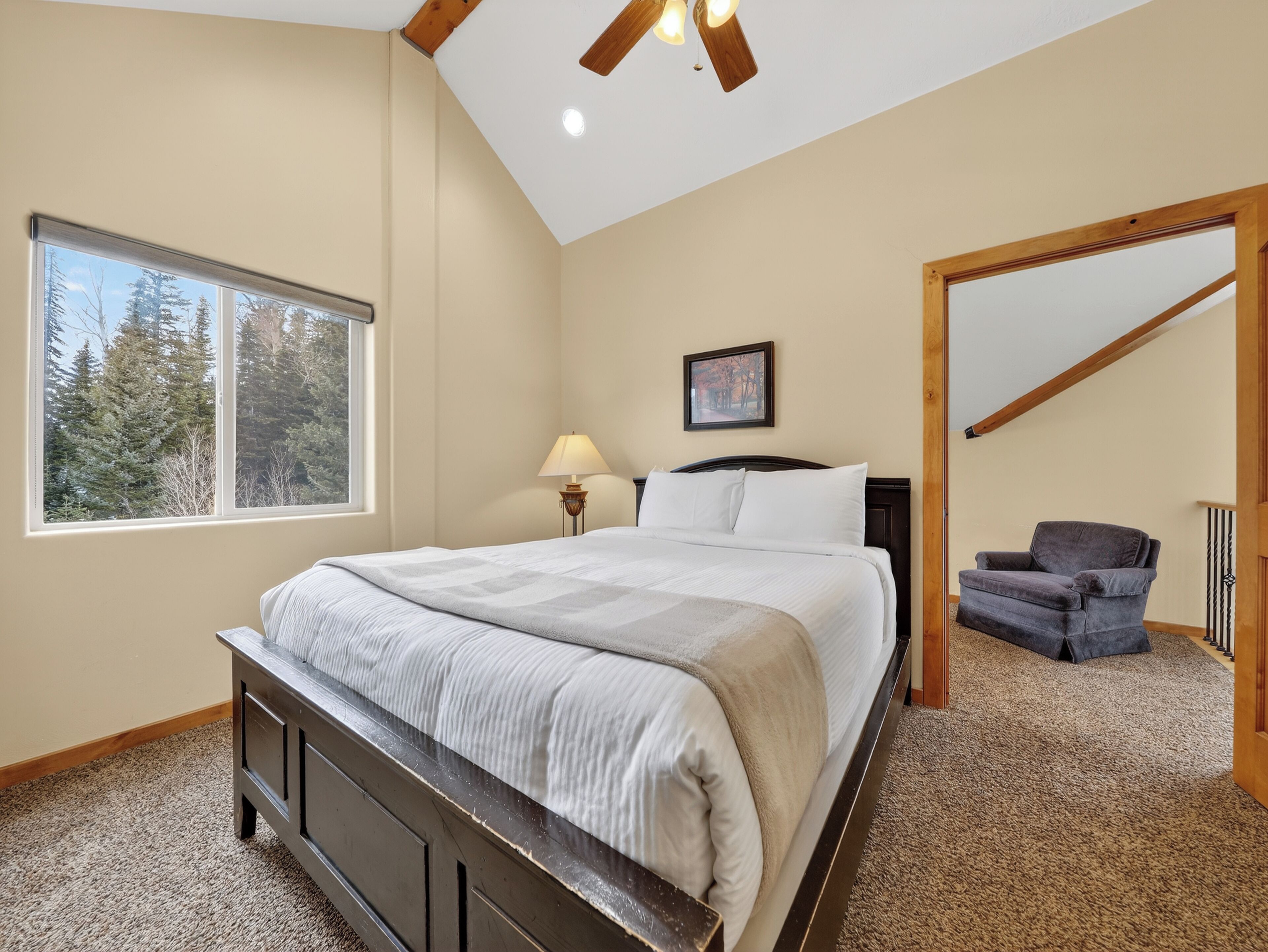 A bedroom with a large bed, a window with a view of trees, a lamp, and a chair.